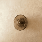 Clérachic flush round wall light with a smoky glass disc and dark bulb in the centre, switched off on a plaster wall.
