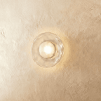 Clérachic flush wall light with a clear rippled glass disc and a frosted bulb glowing warm on a plaster wall.