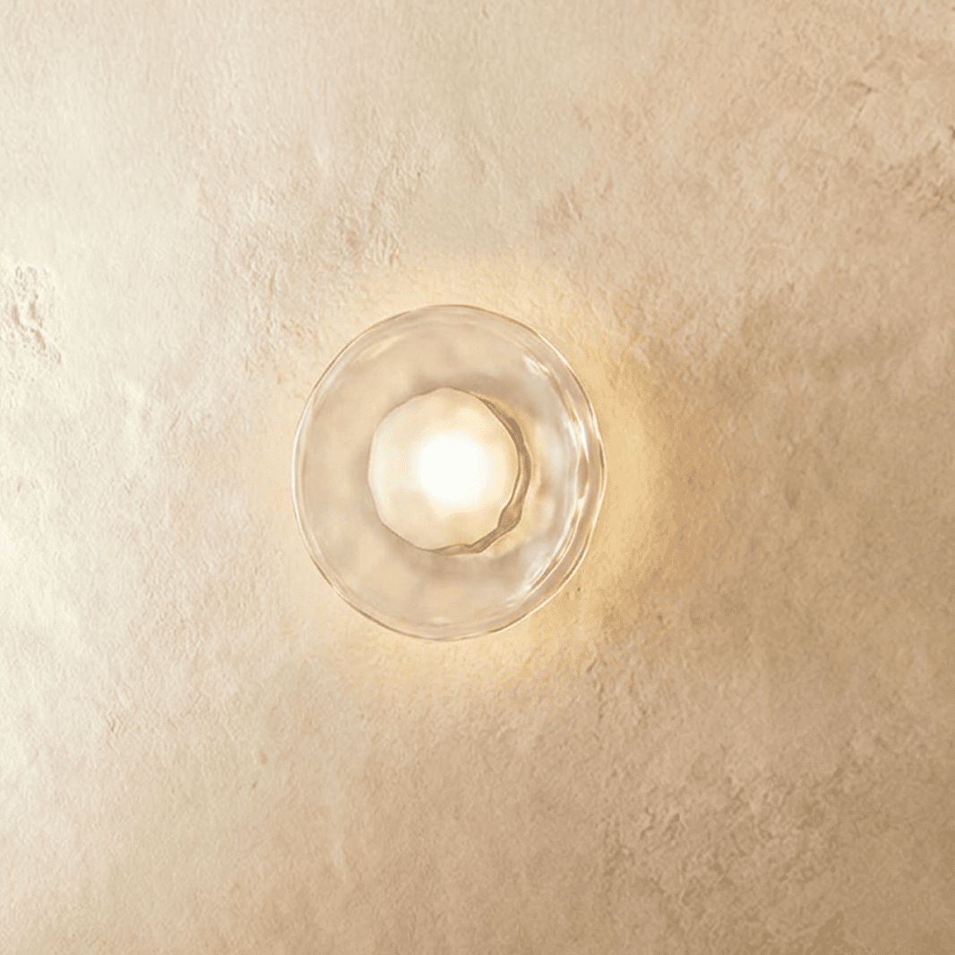 Clérachic flush wall light with a clear rippled glass disc and a frosted bulb glowing warm on a plaster wall.