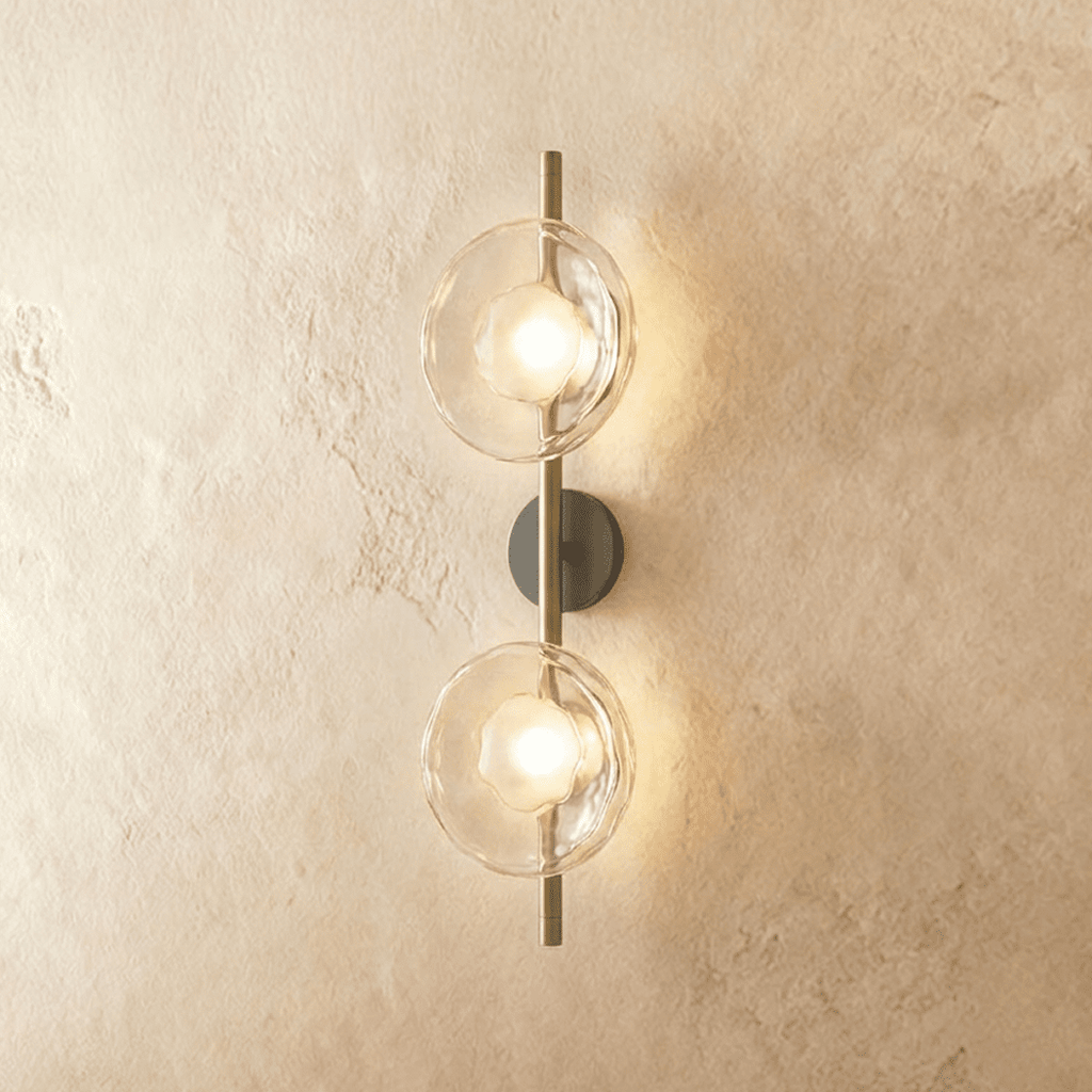 Clérachic two-shade wall light with clear rippled glass discs glowing warm on a brass vertical rod and dark backplate.