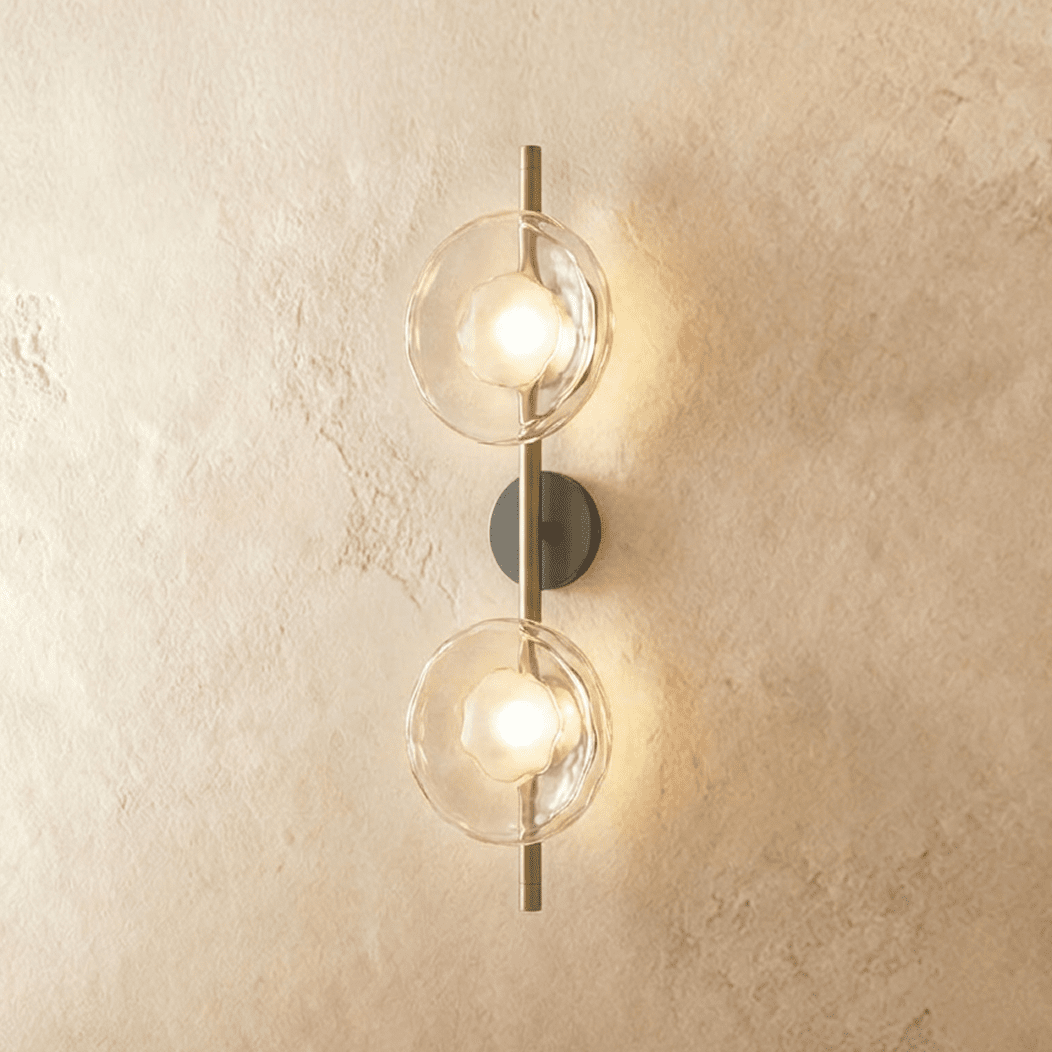 Clérachic two-shade wall light with clear rippled glass discs glowing warm on a brass vertical rod and dark backplate.