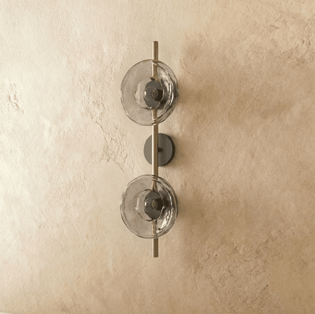 Clérachic two-shade wall light with smoky glass discs on a brushed brass rod and dark backplate, switched off.
