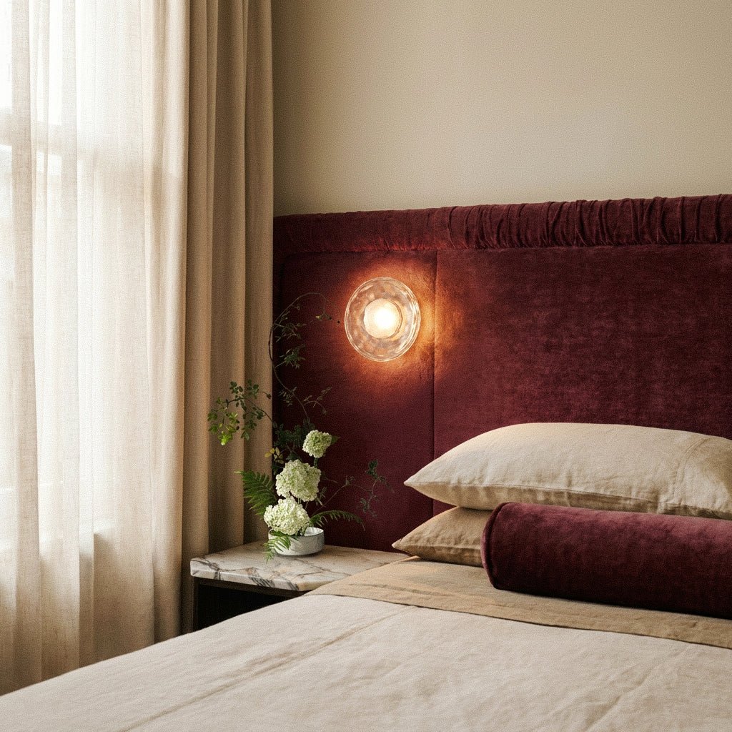 Clérachic round clear glass wall light glowing warm above a bed with a burgundy velvet headboard, linen and white hydrangea.