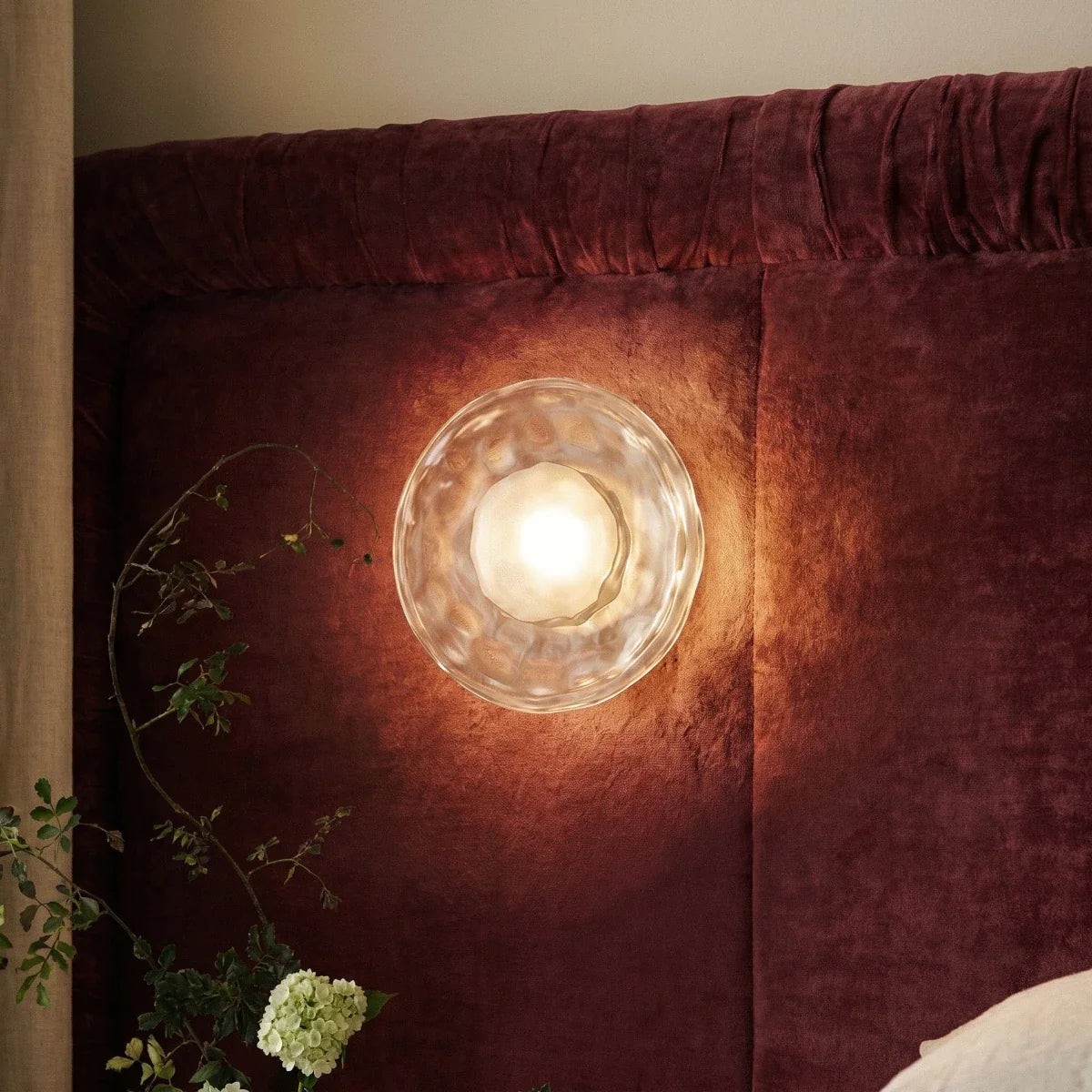 Close-up of a Clérachic round rippled glass wall light with a frosted bulb glowing on a burgundy velvet headboard.
