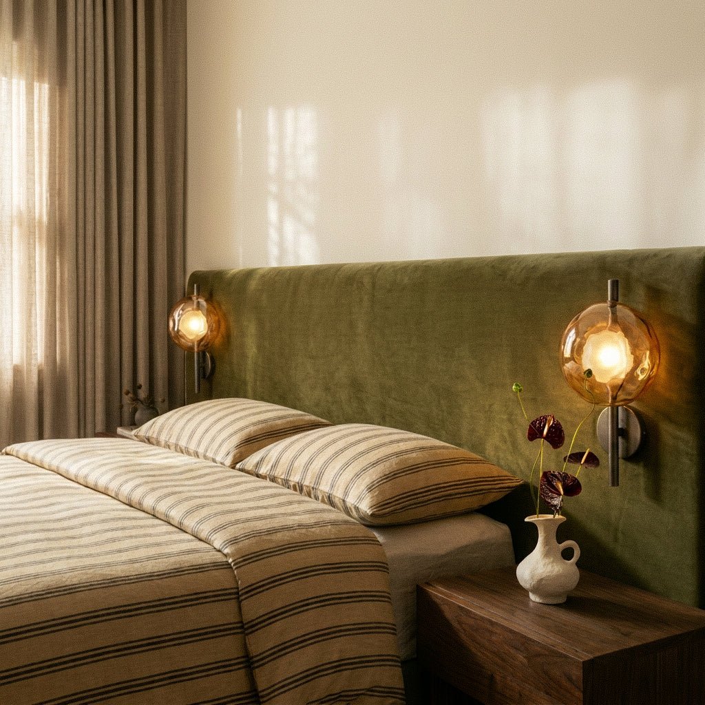 Clérachic pair of amber glass wall lights on black rods glowing either side of a green velvet headboard with striped linen.