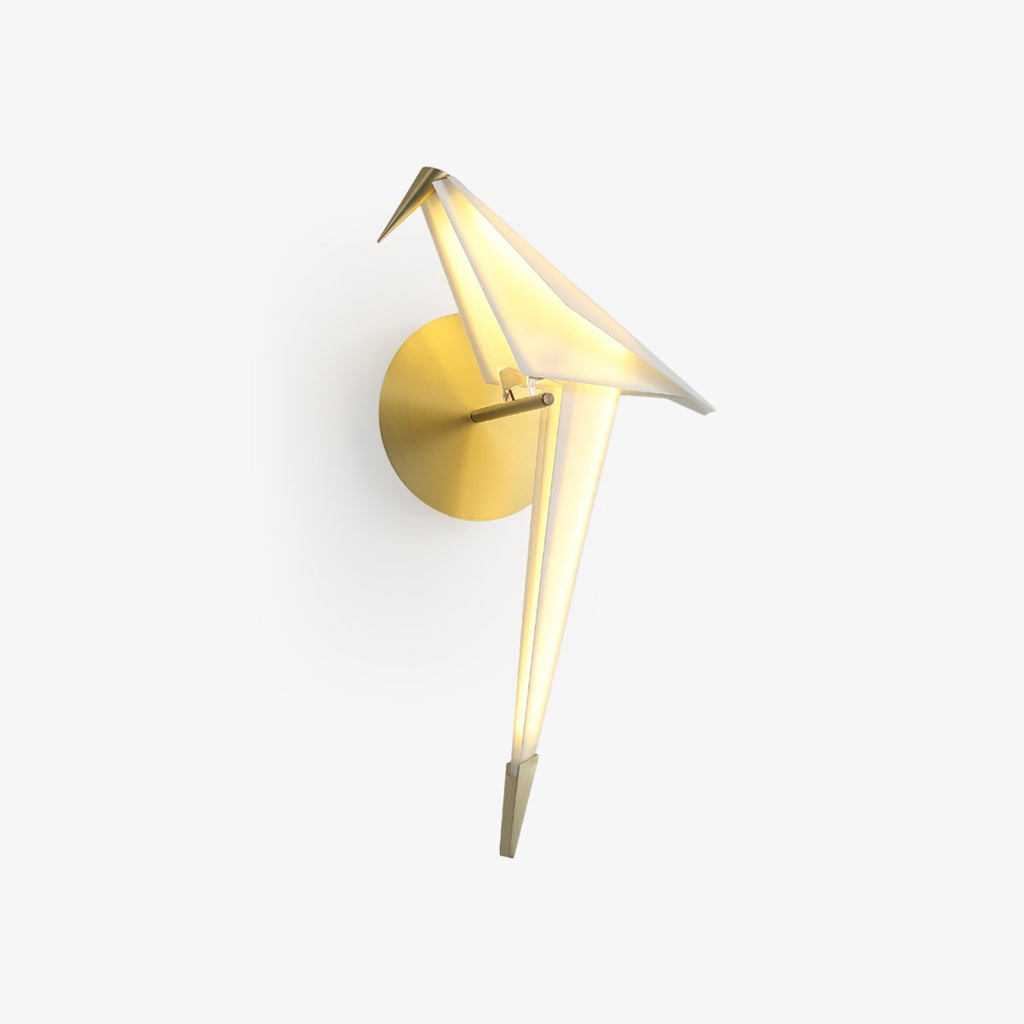 A Clérachic wall light shaped as a folded white bird perched on a gold bar, glowing warm, on a plain white backdrop.