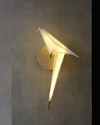 A Clérachic bird wall light glows warm on a textured grey-brown wall, its white body and gold tail tip lit from within.