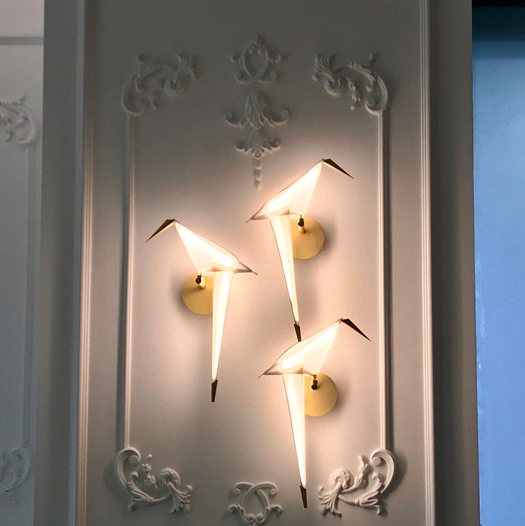 Three Clérachic bird wall lights glow at different heights on an ornate white panelled wall, each on its own round gold plate.