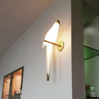 A Clérachic bird wall light mounted high on a white wall by a corner, its white body lit and its gold perch pointing out.