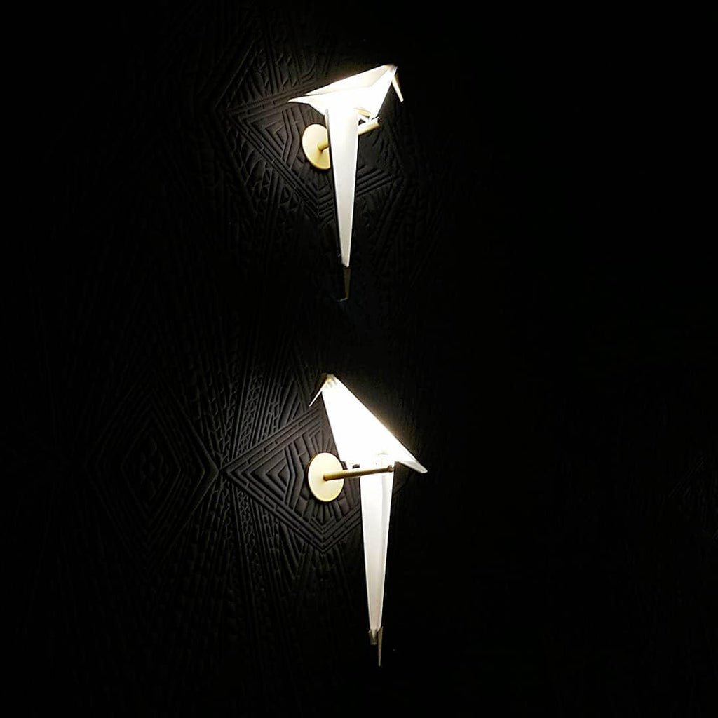 Two Clérachic bird wall lights glow white on a black patterned wall, one above the other, each on a small gold plate.