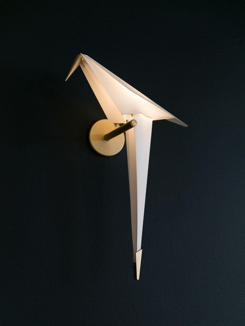 A Clérachic bird wall light, unlit, against a deep navy wall, its folded white body and gold perch standing out sharply.