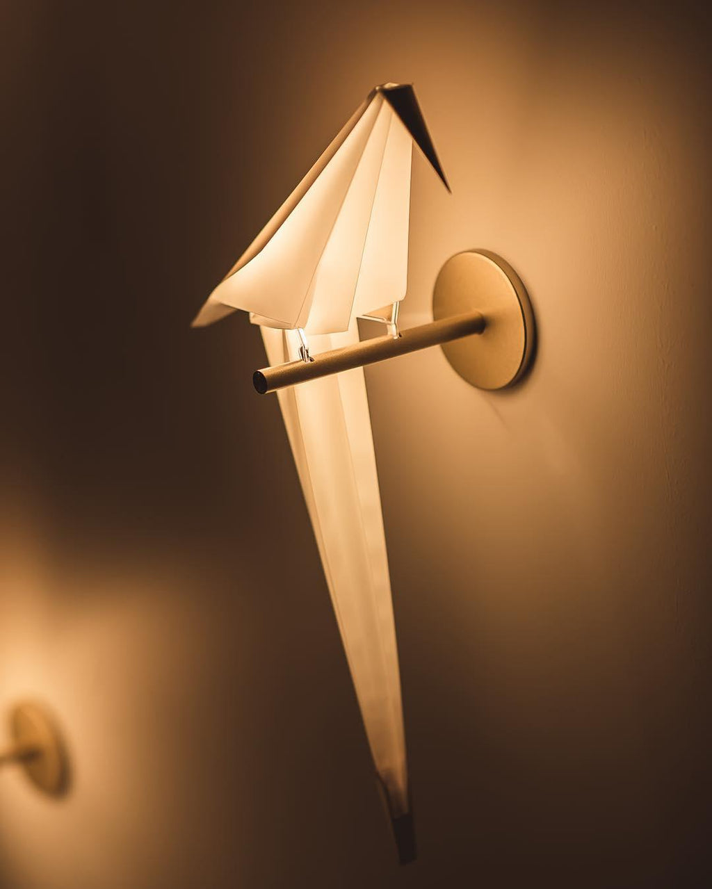 Close view of a lit Clérachic bird wall light on a warm brown wall, its glowing white tail running down to a gold tip.