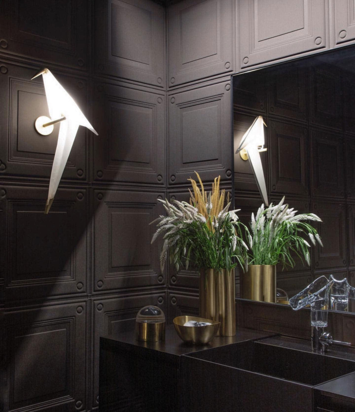 A Clérachic bird wall light glows on a dark panelled wall above a black counter, with a mirror and gold vases of grasses below.