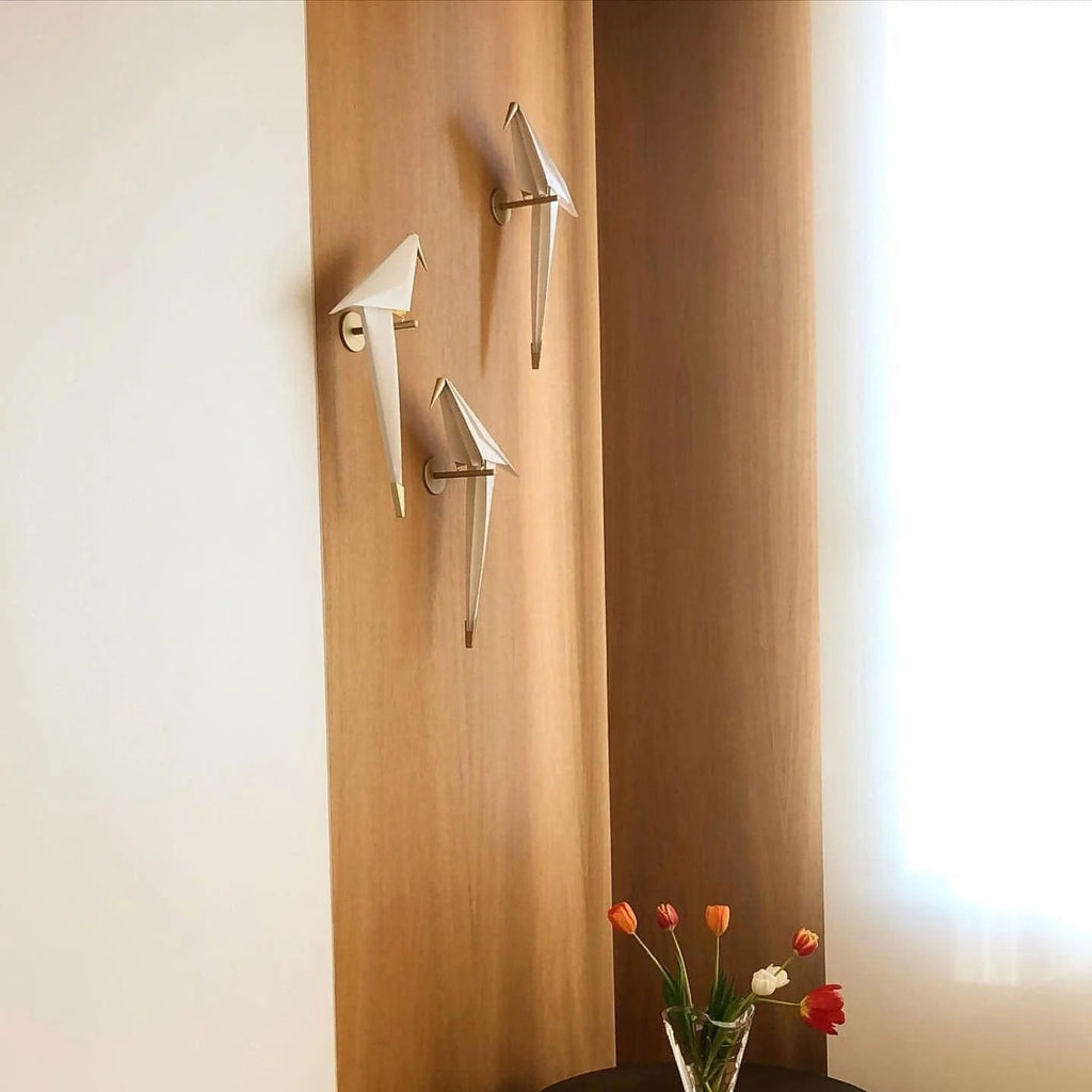 Three Clérachic bird wall lights on a pale timber panel by a window, unlit, with a glass vase of tulips below.