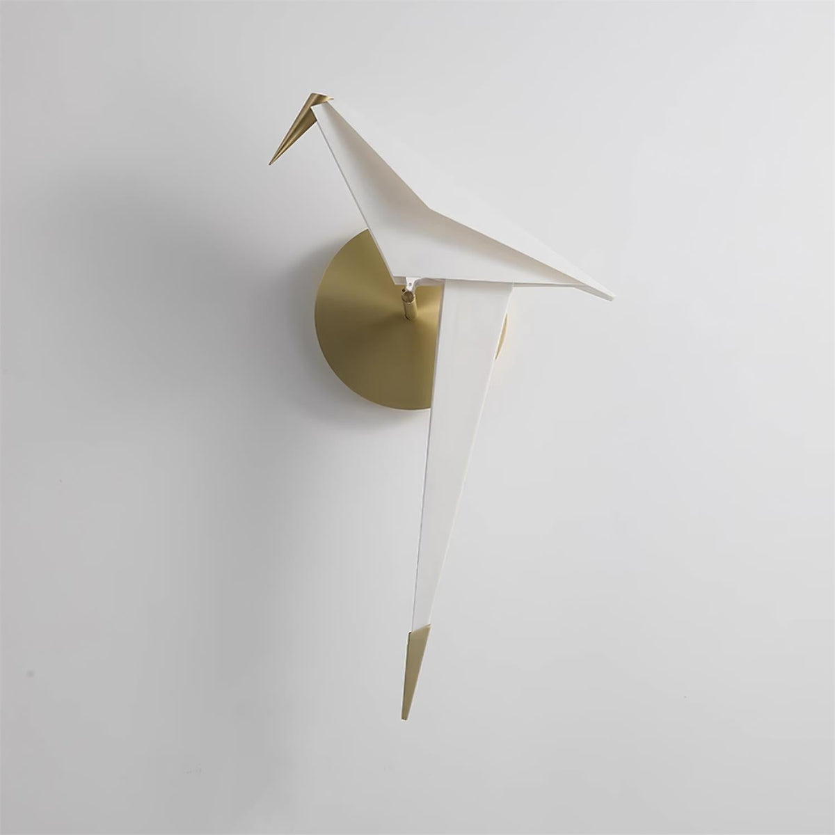 A Clérachic bird wall light, unlit, on a pale wall, its folded white body, gold tail tip and brushed gold backplate in view.