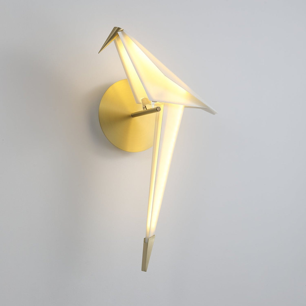 A lit Clérachic bird wall light on a grey wall, its folded white body glowing along the tail above a brushed gold backplate.