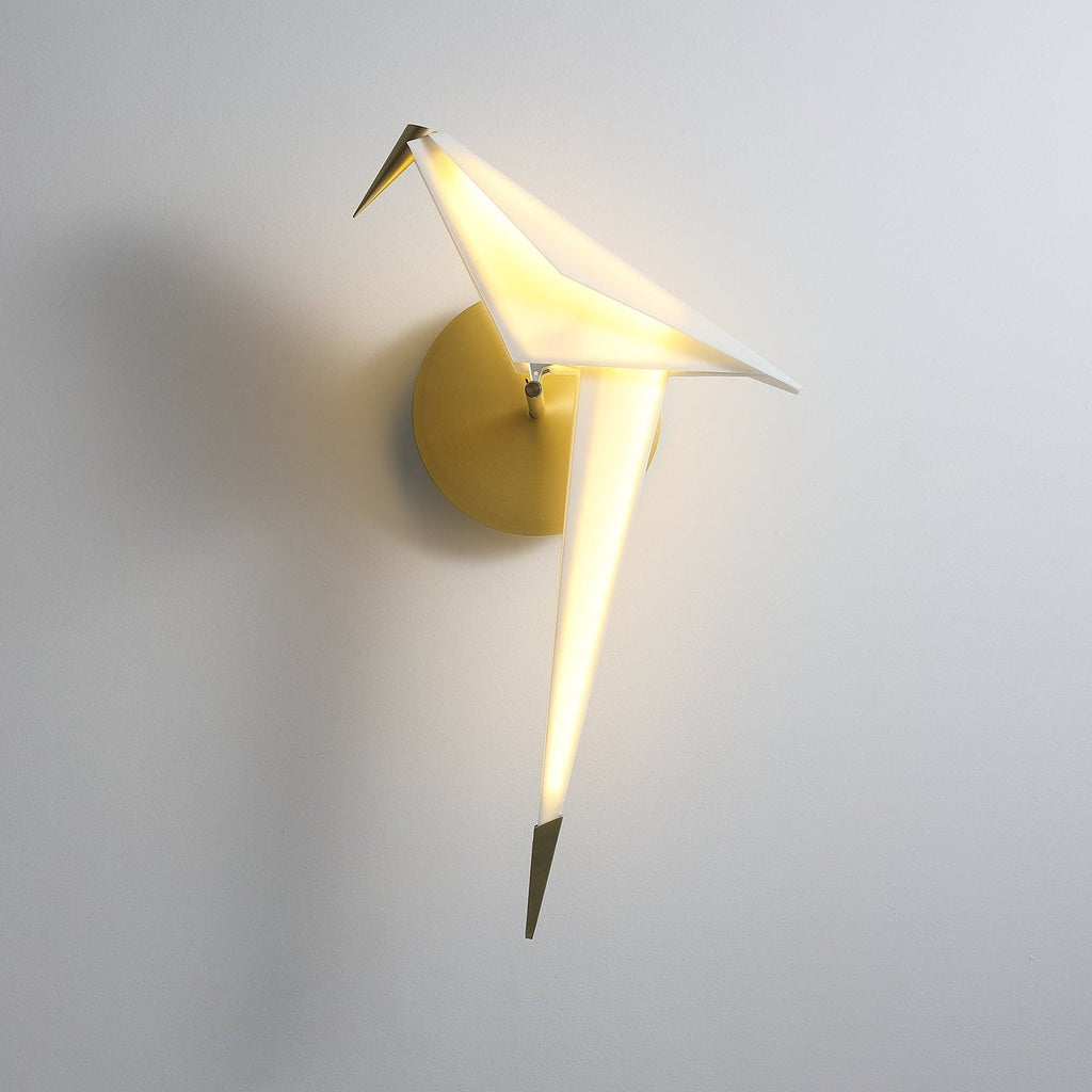 Front view of a lit Clérachic bird wall light on a pale wall, its white body glowing and its gold tail pointing down.