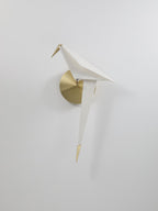 A smaller view of this unlit Clérachic bird wall light on a pale wall, with a brushed gold backplate and a gold tail tip.