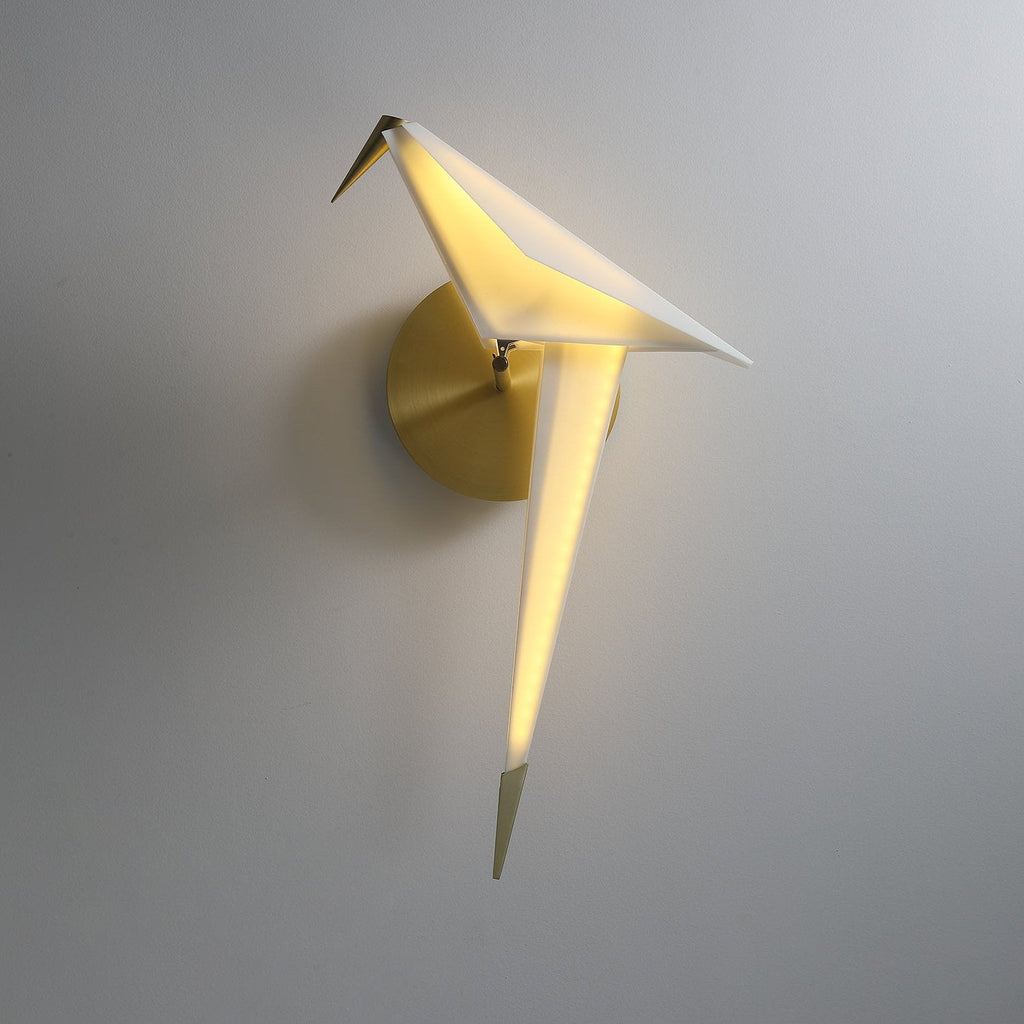 A lit Clérachic bird wall light on a grey wall, with a soft glow along its white tail and a gold backplate behind its perch.