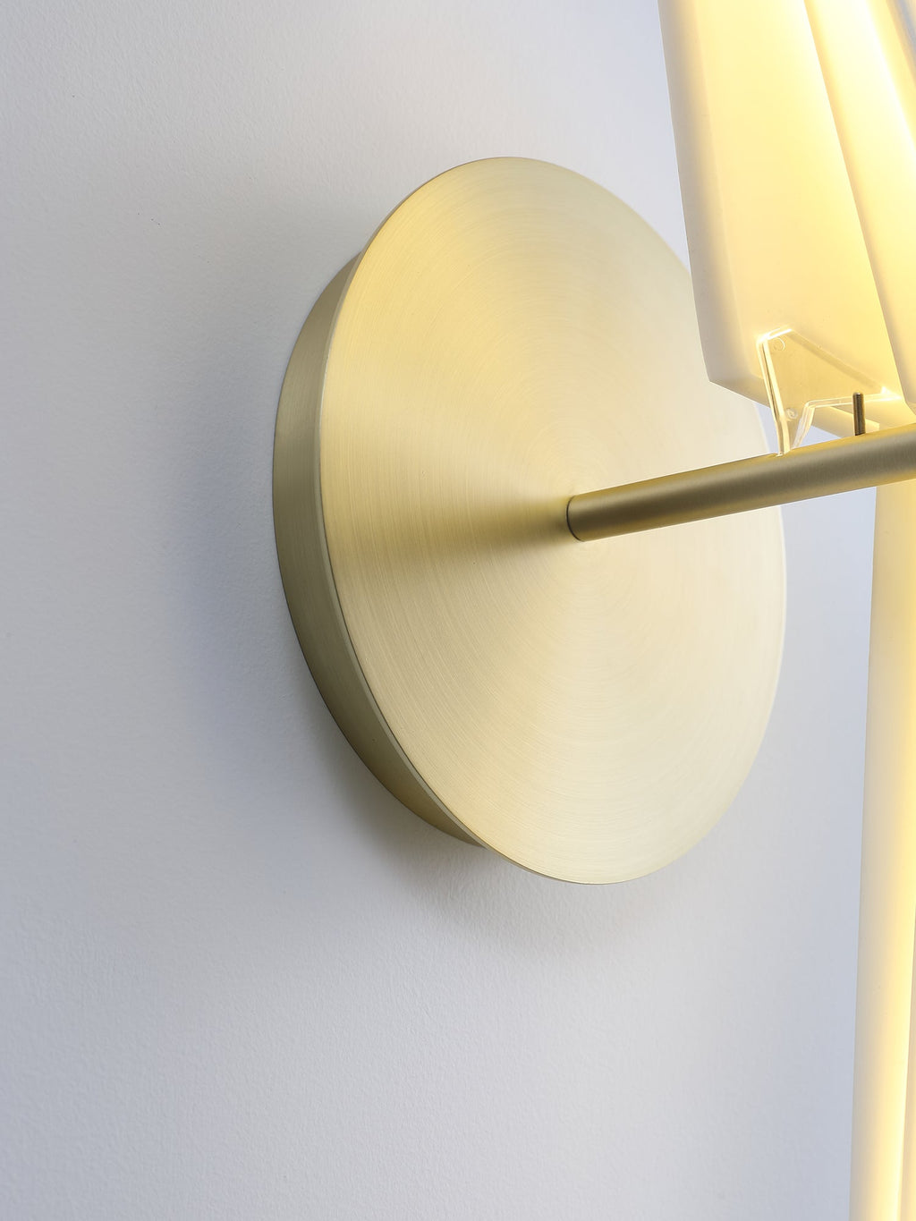 Seen from the side, the brushed gold round backplate and the perch bar of a Clérachic bird wall light, with the lit body at the edge.