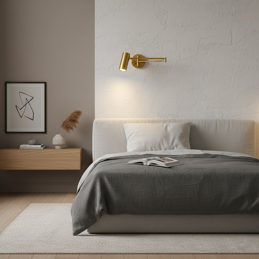 A Clérachic gold wall light above a bed with grey bedding, on a textured plaster wall beside a framed print.
