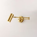Clérachic gold wall light with its head tilted down, on a plain pale wall, with the arm folded at the elbow.