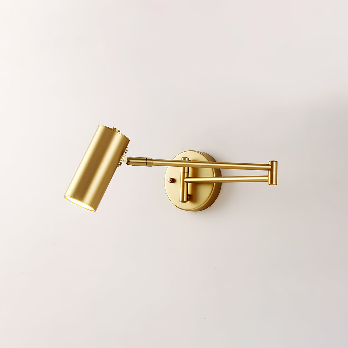 Clérachic gold wall light with its head tilted down, on a plain pale wall, with the arm folded at the elbow.