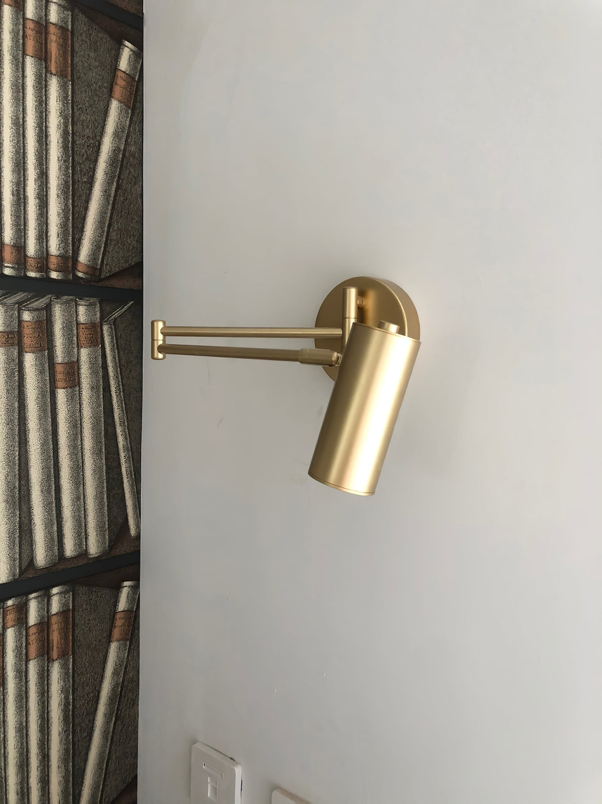 A Clérachic gold wall light with a folded arm, mounted on a plain wall beside a bookcase-patterned panel.