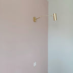 A small Clérachic gold wall light with a long extended arm, seen from far away on a blush-pink wall.
