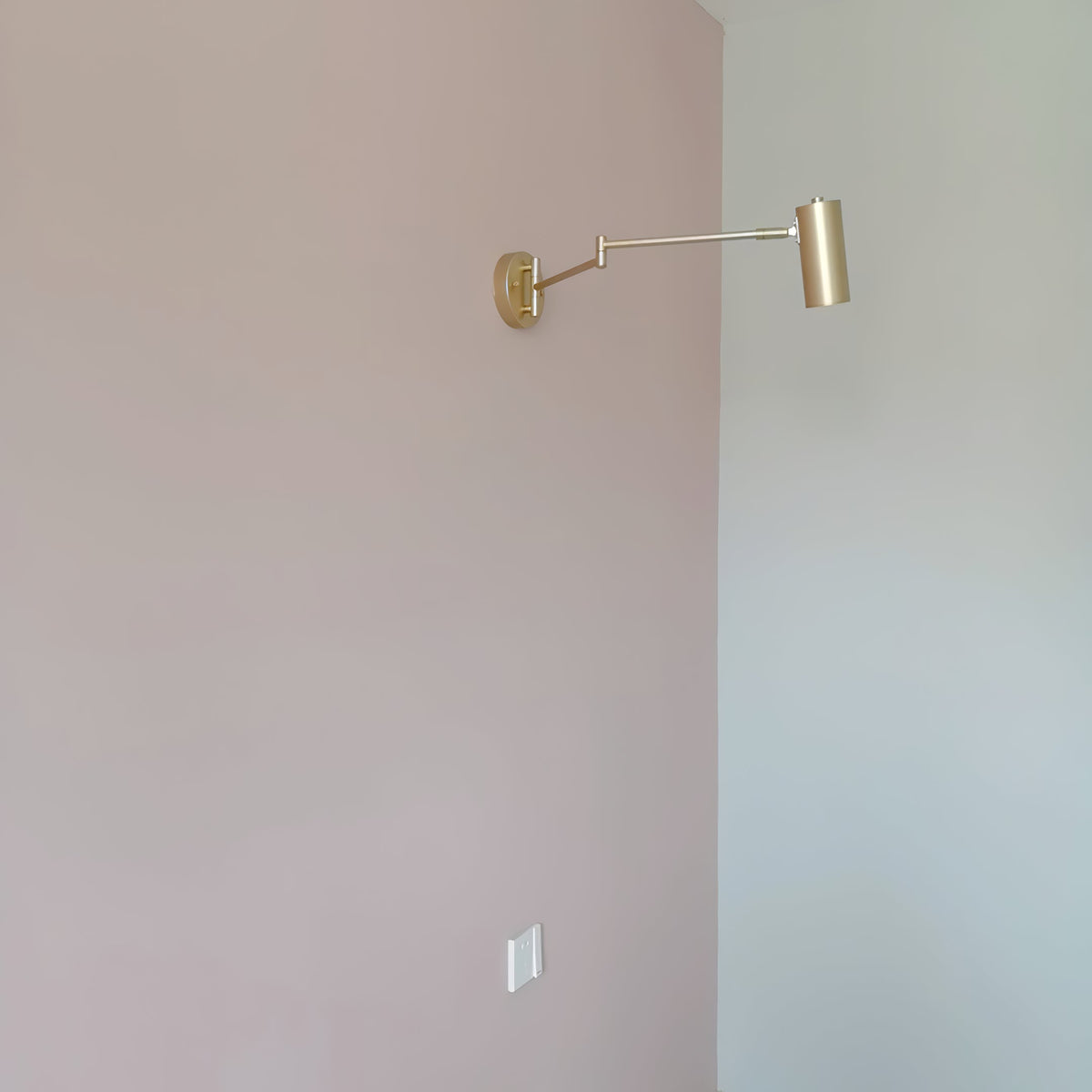 A small Clérachic gold wall light with a long extended arm, seen from far away on a blush-pink wall.