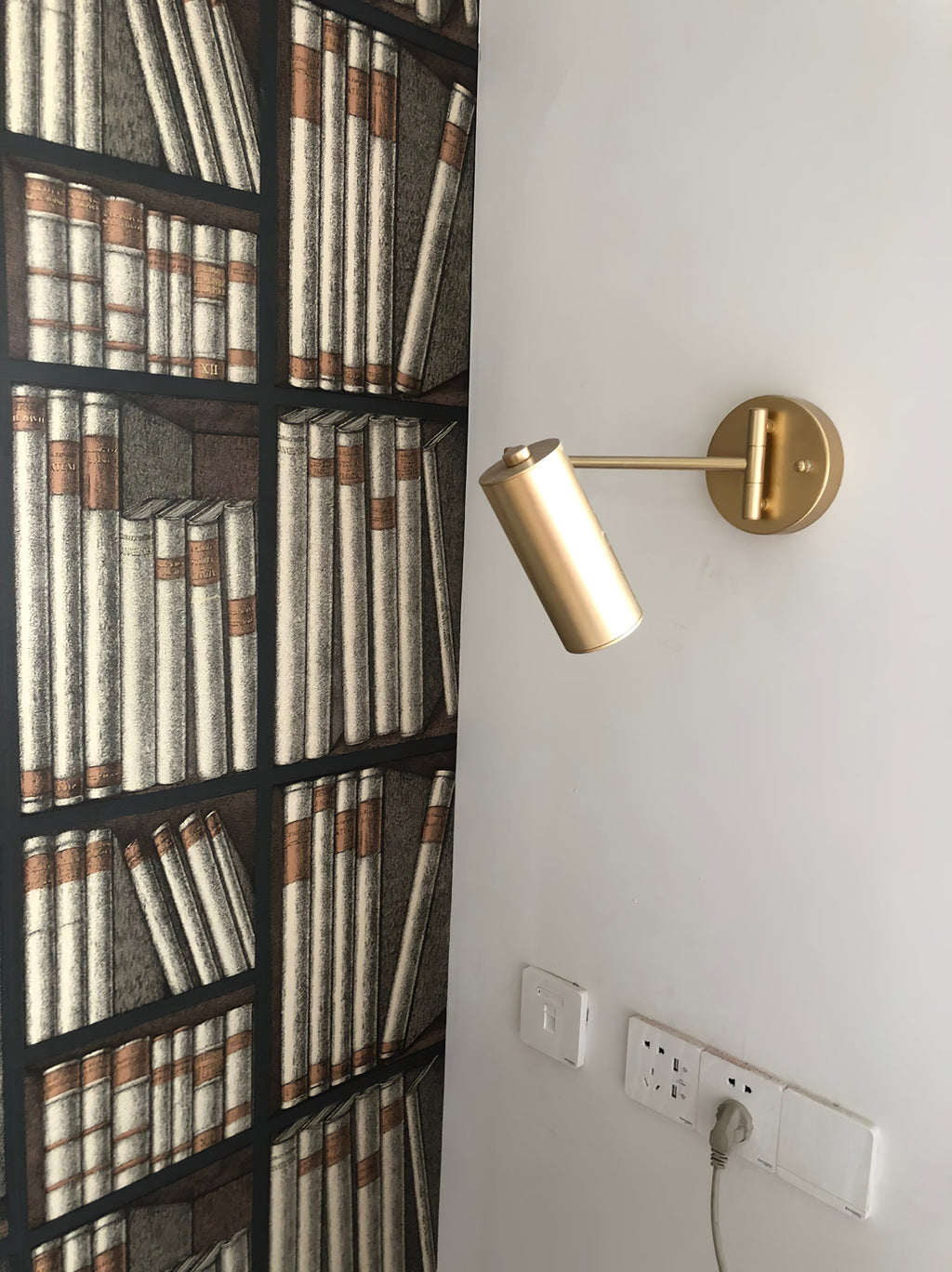 Clérachic gold wall light mounted beside a bookcase-patterned panel, above a power socket strip.