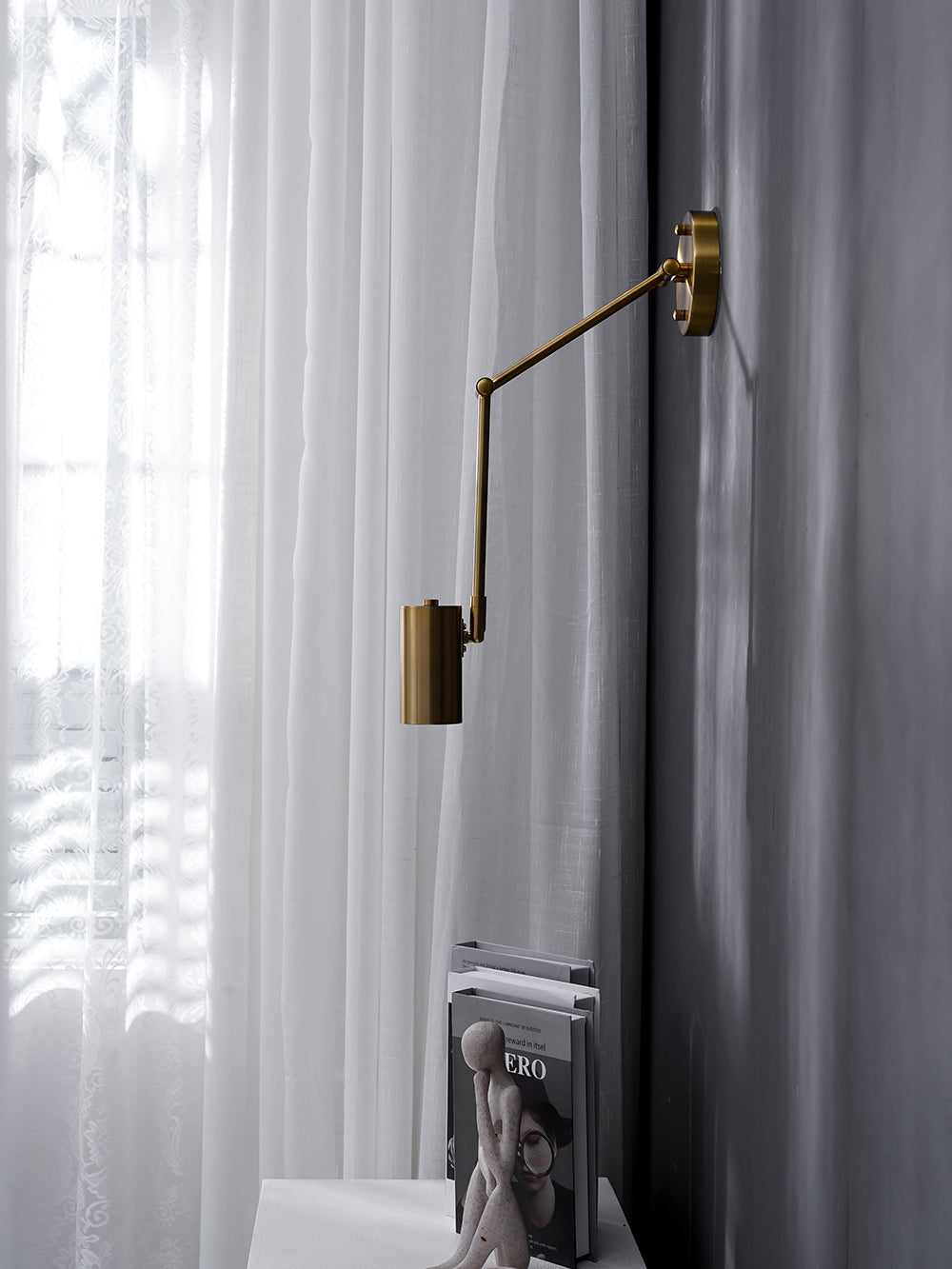 Clérachic gold wall light with its drop arm, hanging above a white console, a stack of books and a sheer curtain.