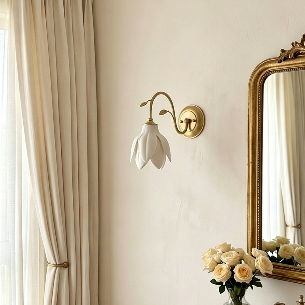 Unlit Clérachic wall light with a white petal shade on a cream wall, beside a gold-framed mirror, sheer curtains and a vase of cream roses.