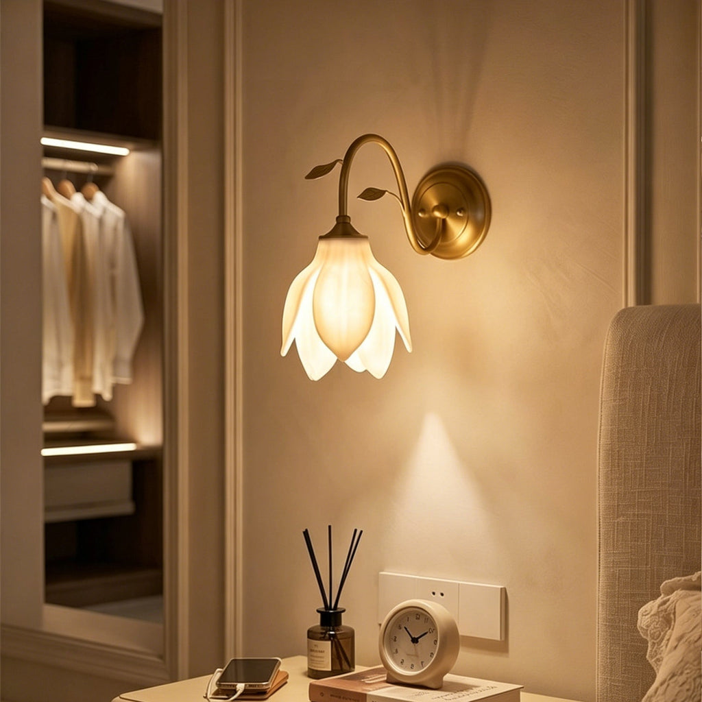 A Clérachic wall light with a glowing white petal shade above a bedside table with a reed diffuser and clock, with a dressing area behind.