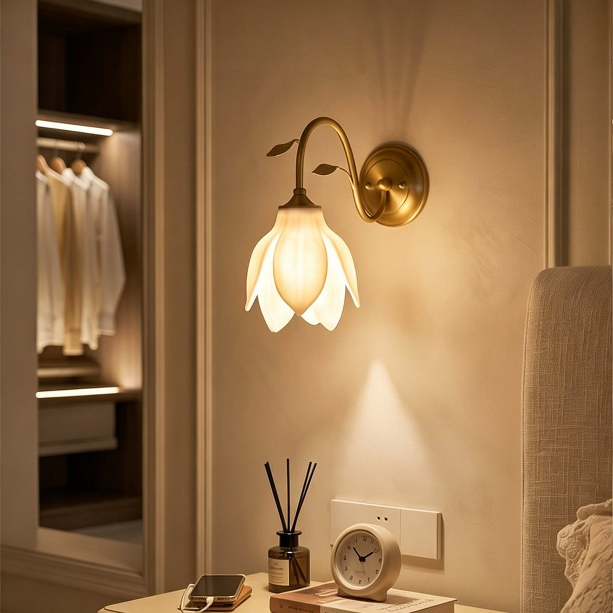 A Clérachic wall light with a glowing white petal shade above a bedside table with a reed diffuser and clock, with a dressing area behind.