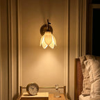 Clérachic wall light with a lit petal shade centred above a small bedside table holding a round clock and a glass perfume bottle.
