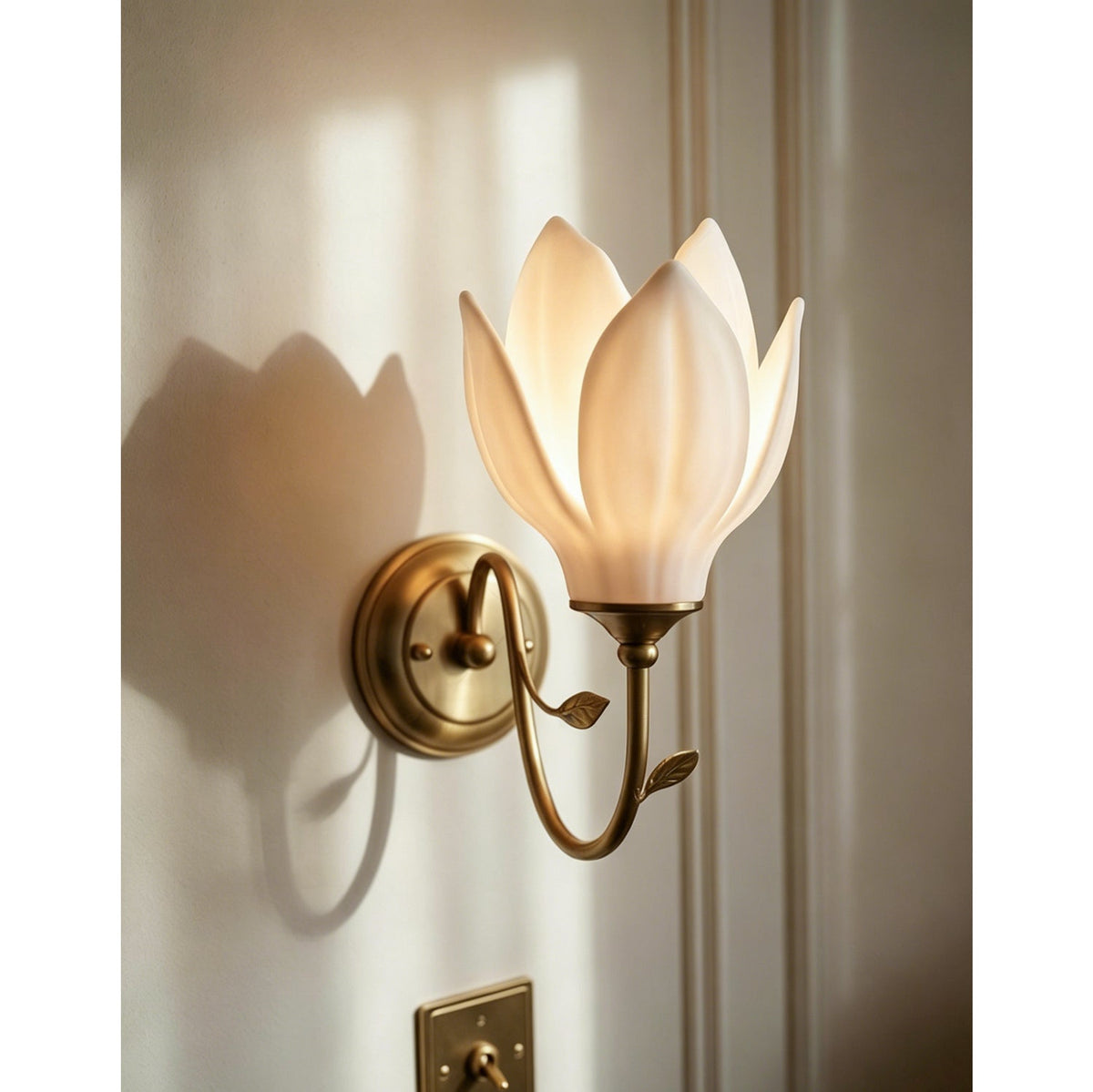 Petal shade pointing up on a gold arm, this Clérachic wall light is lit against a pale wall with a soft shadow and a brass switch below.