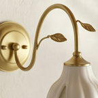 Close view of the round gold wall plate, curved arm and two leaf details above the white ceramic shade of this Clérachic wall light.