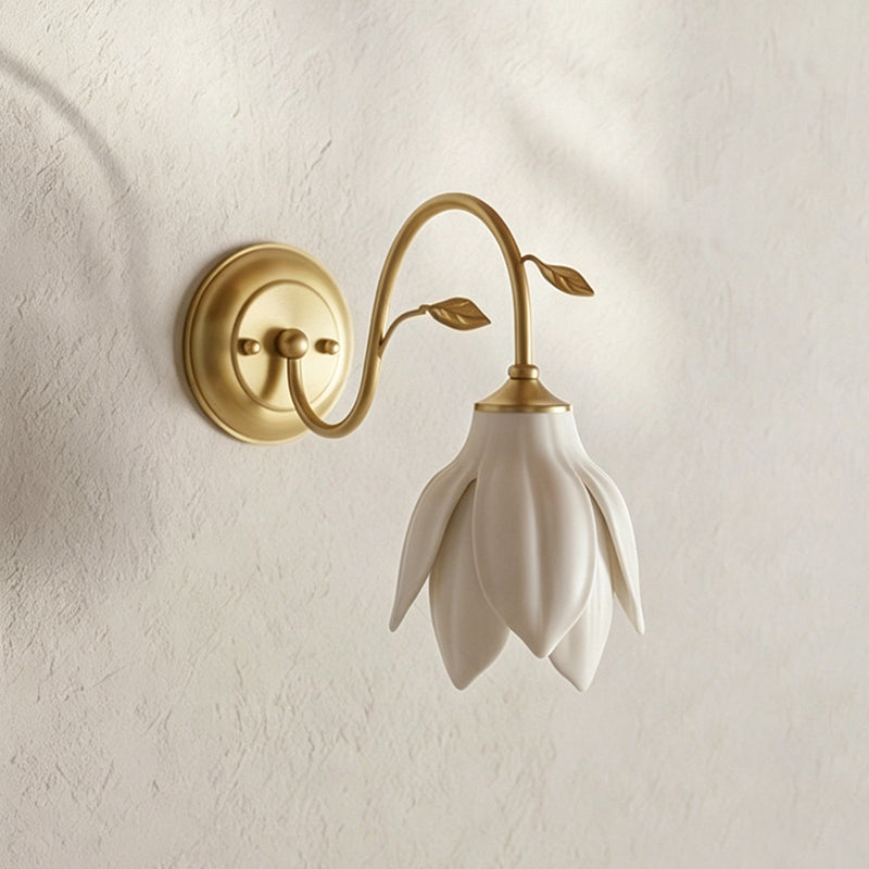 This Clérachic wall light hangs its white ceramic petal shade from a gold arm against a textured cream wall with soft shadows.