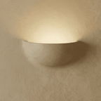 Clérachic half-moon plaster wall light glowing upward on a limewashed wall, its matte white shell casting a warm halo.