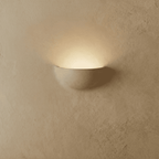 Clérachic half-moon plaster wall light in matte white lit from within, throwing a soft arc of light up a textured wall.