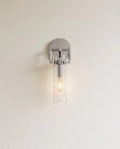 Clérachic polished chrome wall sconce with a clear glass cylinder shade and a warm filament bulb on a pale wall.