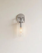 Clérachic polished chrome wall sconce with a clear glass cylinder shade and a warm filament bulb on a pale wall.