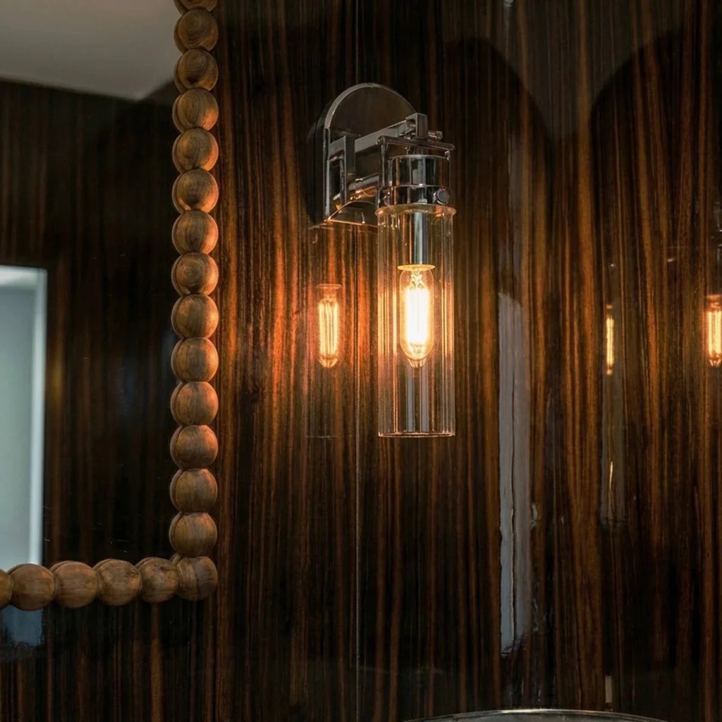 Clérachic chrome glass-cylinder sconce glowing amber on a dark striped wood wall beside a beaded wooden mirror frame.