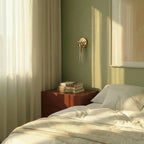 Clérachic gold glass-cylinder sconce on a sage green wall above a rust-red bedside table with stacked books.