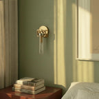 Clérachic gold sconce with a clear glass cylinder shade on a sage green wall, above a bedside table with books.