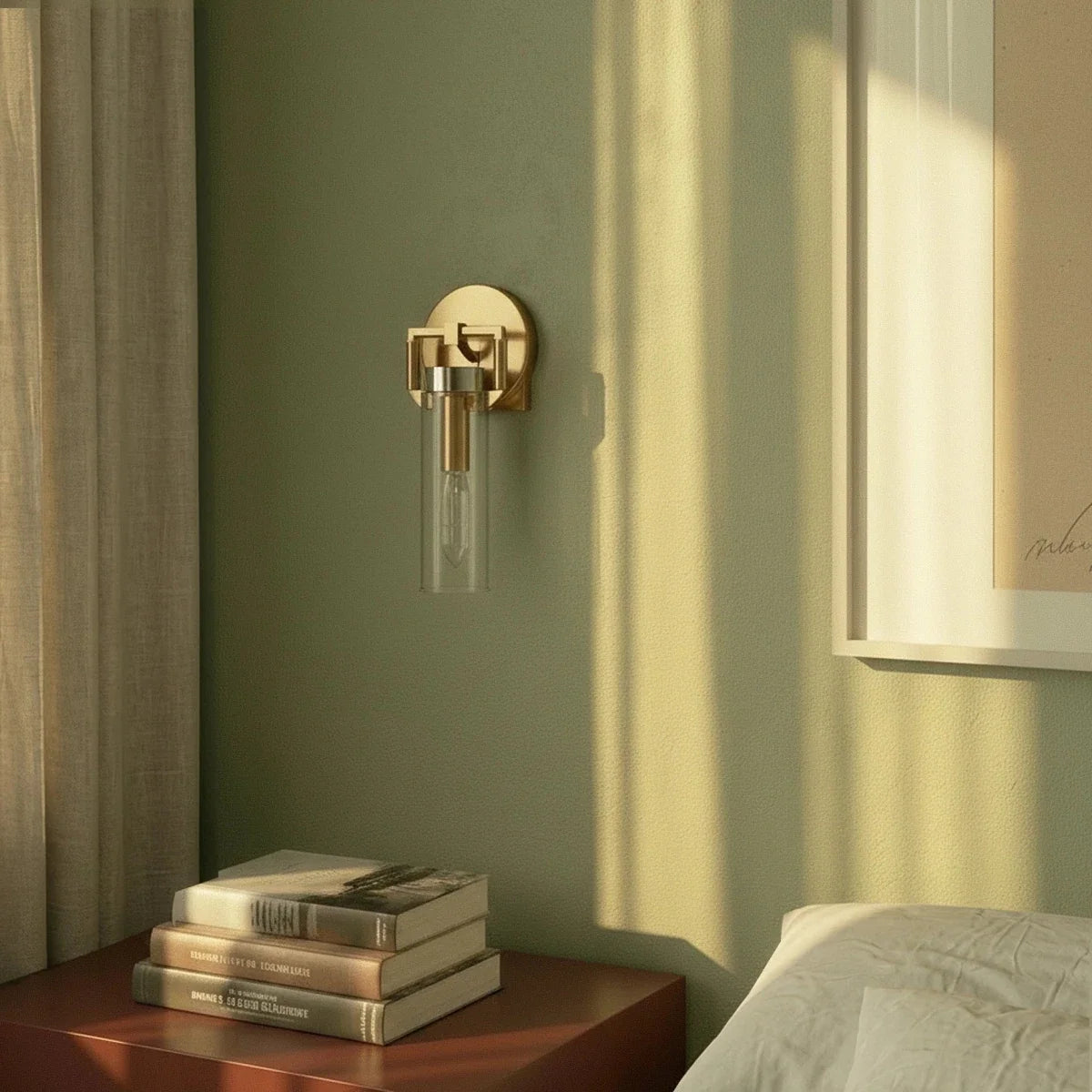 Clérachic gold sconce with a clear glass cylinder shade on a sage green wall, above a bedside table with books.
