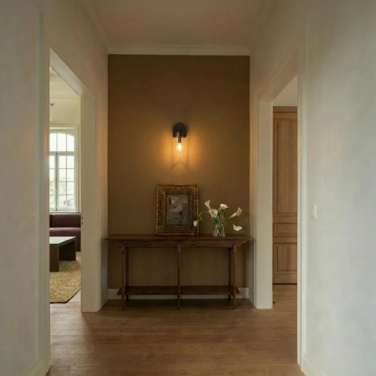 Clérachic black glass-cylinder sconce lit above a console table with a framed print and white lilies in a hallway.
