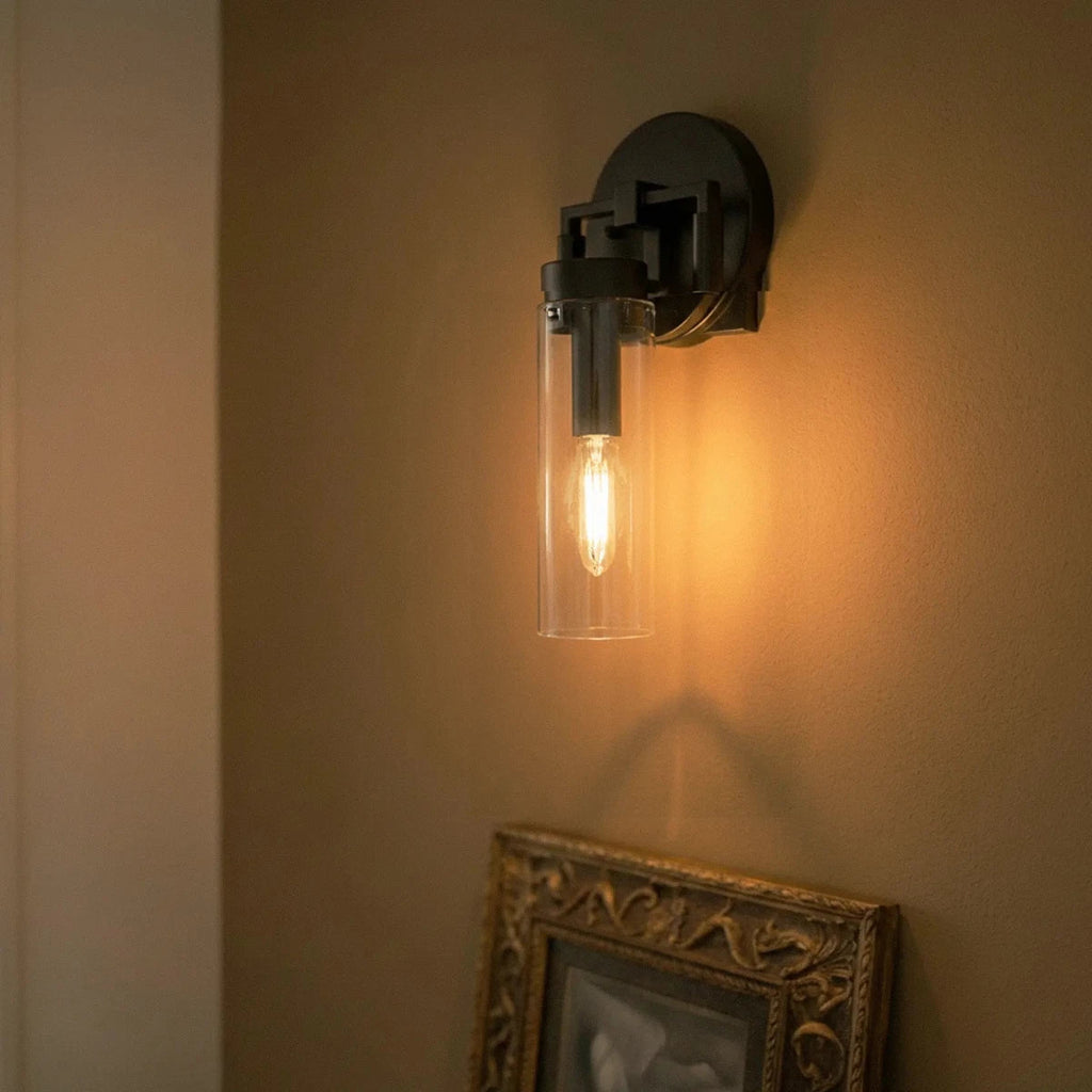 Clérachic black sconce glowing amber on a warm wall above an ornate gilt-framed picture, a filament bulb inside the glass.