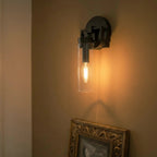 Clérachic black sconce glowing amber on a warm wall above an ornate gilt-framed picture, a filament bulb inside the glass.